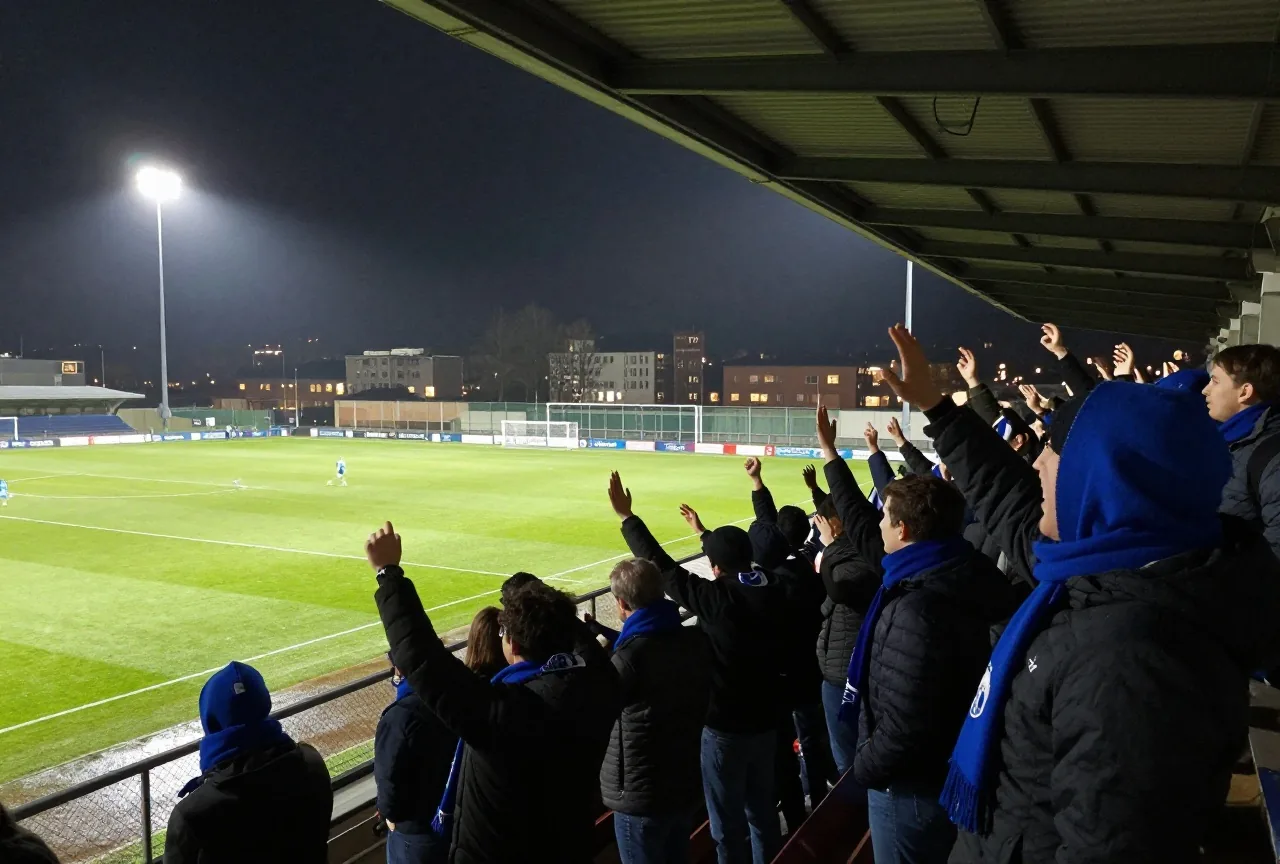 Fußballfans mit blauen Schals jubeln nachts auf einer überdachten Stehtribüne im Flutlicht