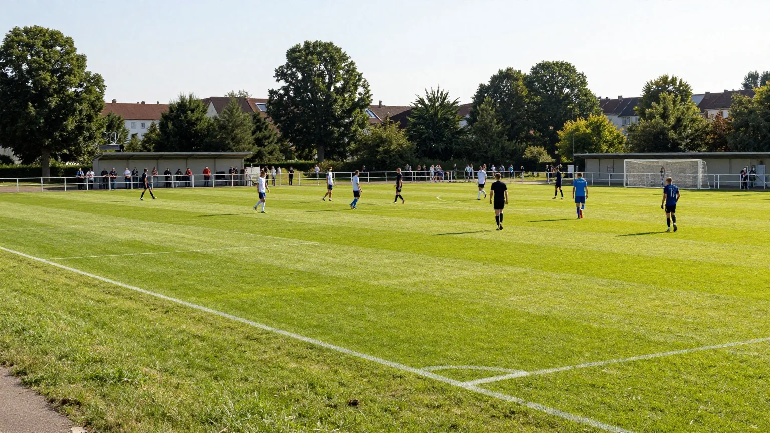 Amateursportplatz am Sonntagnachmittag mit Spielern, Zuschauern am Geländer und kleinem Vereinsheim