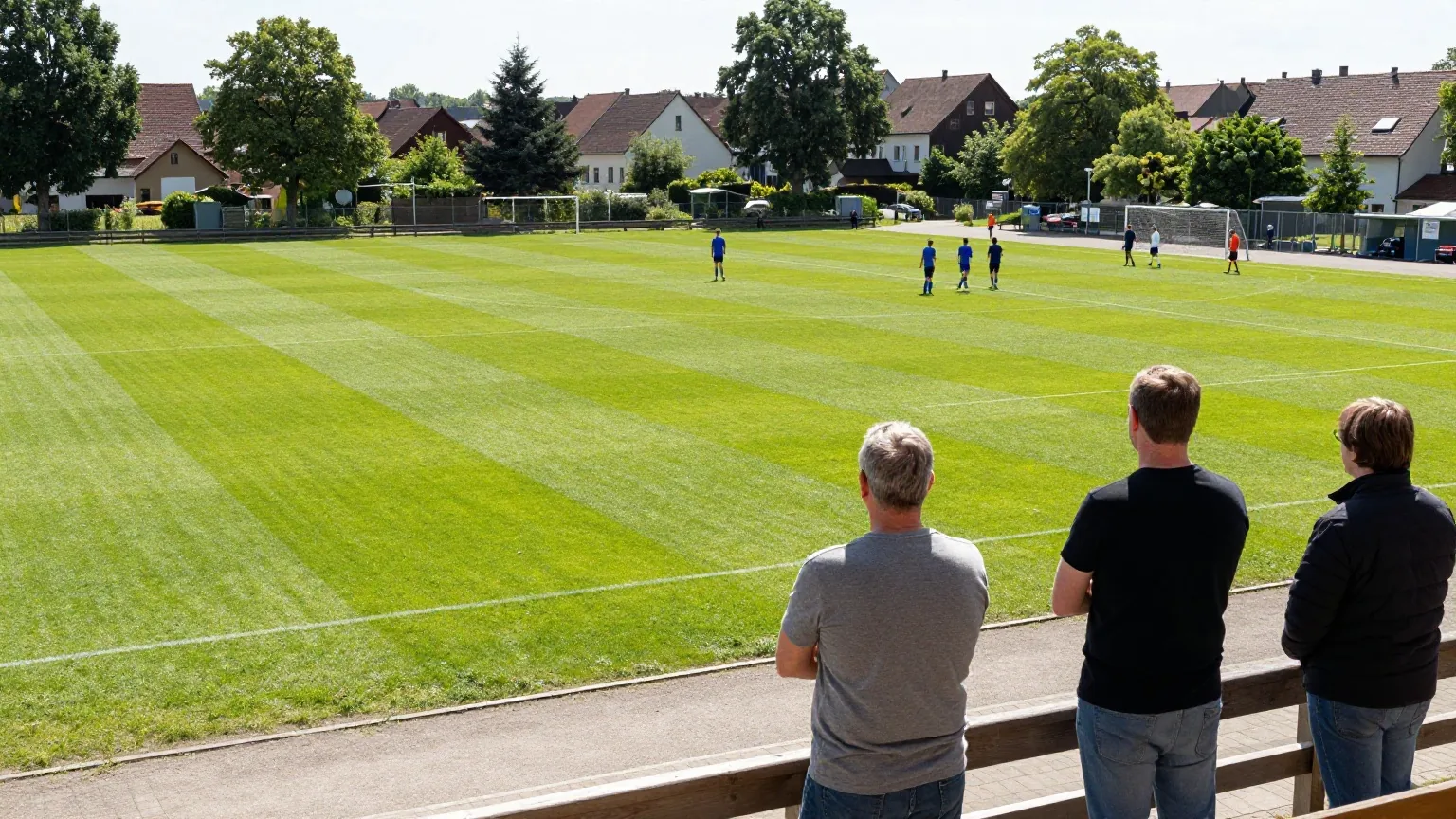 Kleiner Dorfsportplatz am Sonntagnachmittag mit Zuschauern am Geländer und Spielern in der Ferne