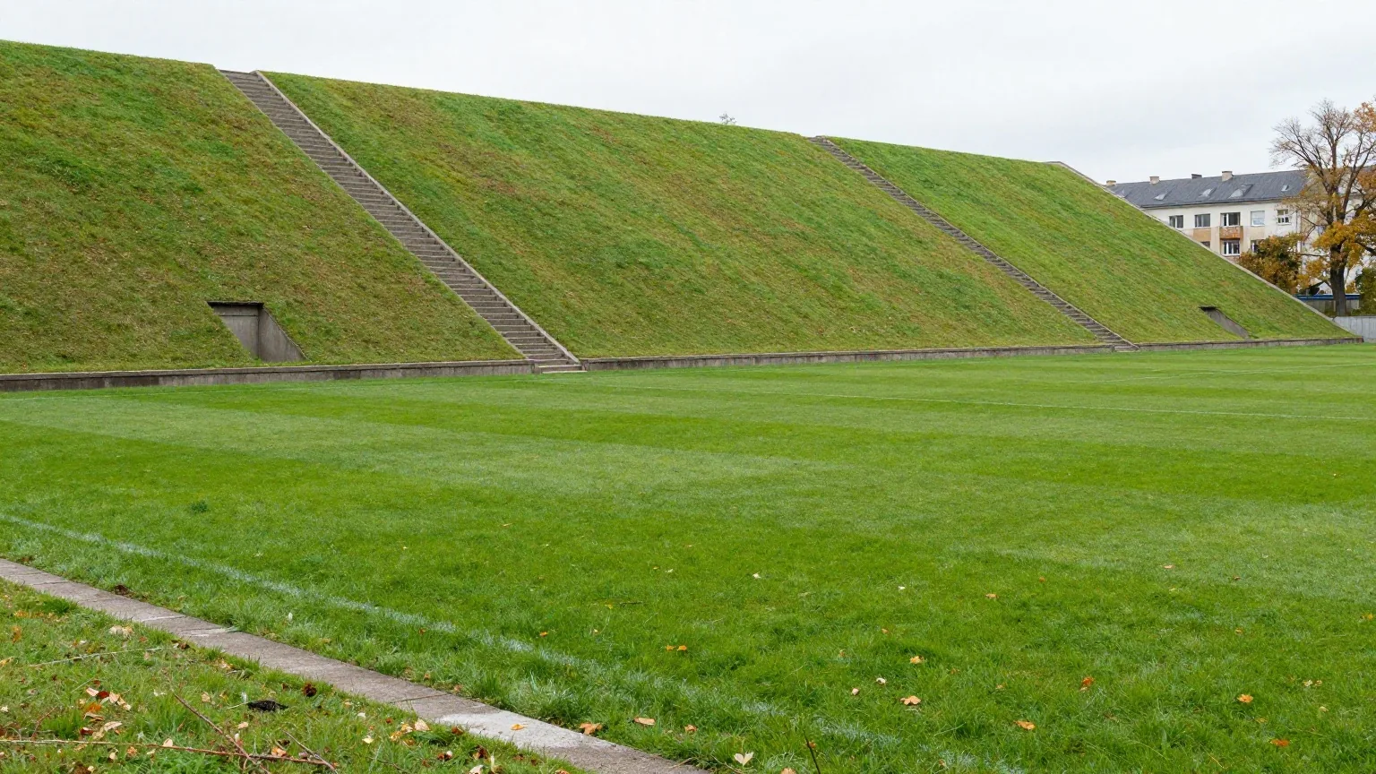 Alter Fußballplatz mit grasbewachsenem Erdwall als Tribüne und schlichten Betonstufen an einem grauen Herbsttag