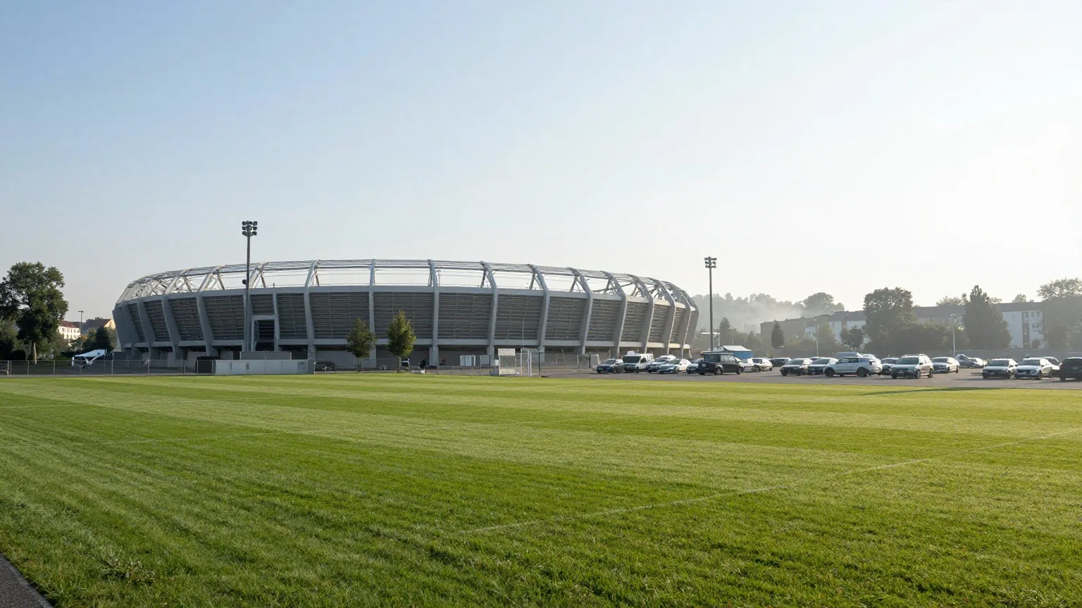 Modernes Fußballstadion am Stadtrand zwischen Bäumen und einem großen Parkplatz im Morgennebel bei tief stehender Sonne