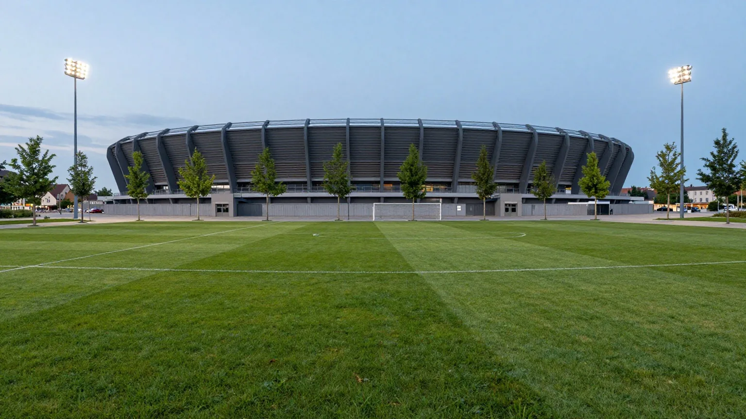 Modernes mittelgroßes Fußballstadion mit dunkelgrauer Fassade zur blauen Stunde, davor ein leerer Platz mit jungen Bäumen
