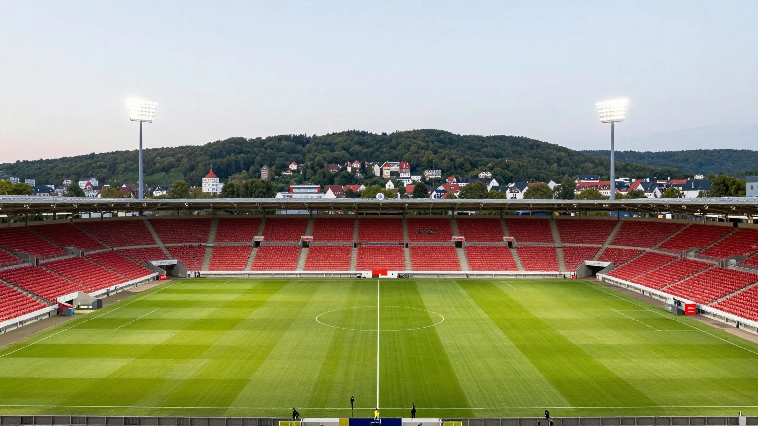 Großes Fußballstadion auf einem bewaldeten Hügel über einer Stadt in der Dämmerung, Flutlicht an