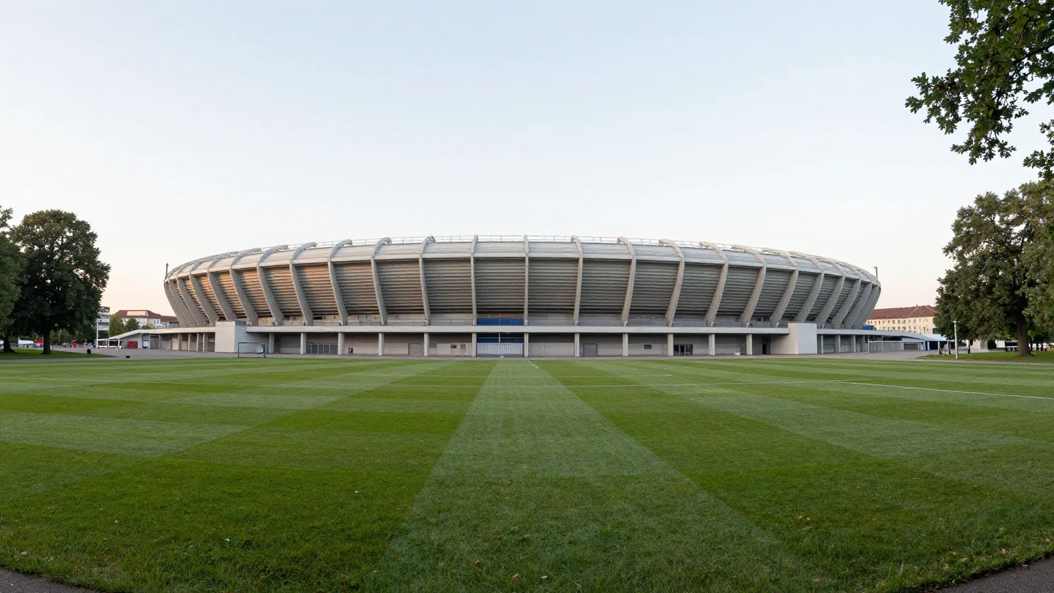 Großes Fußballstadion mit historischer Haupttribüne im Bauhausstil neben einem Park mit alten Bäumen im Morgenlicht