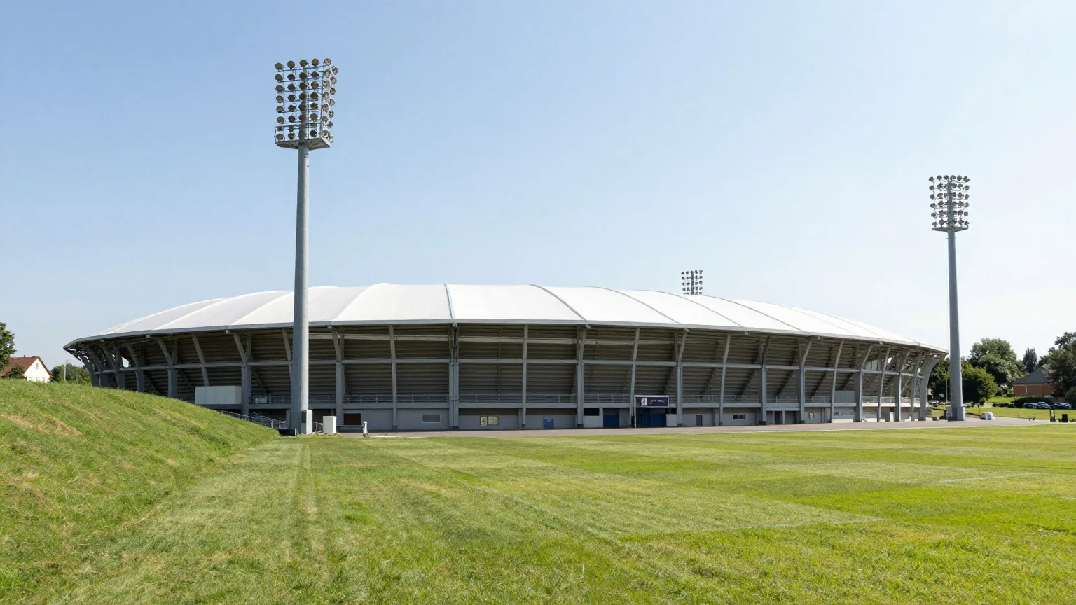 Kompaktes Fußballstadion mit weißen Membrandächern und hohen alten Flutlichtmasten an einem sonnigen Nachmittag