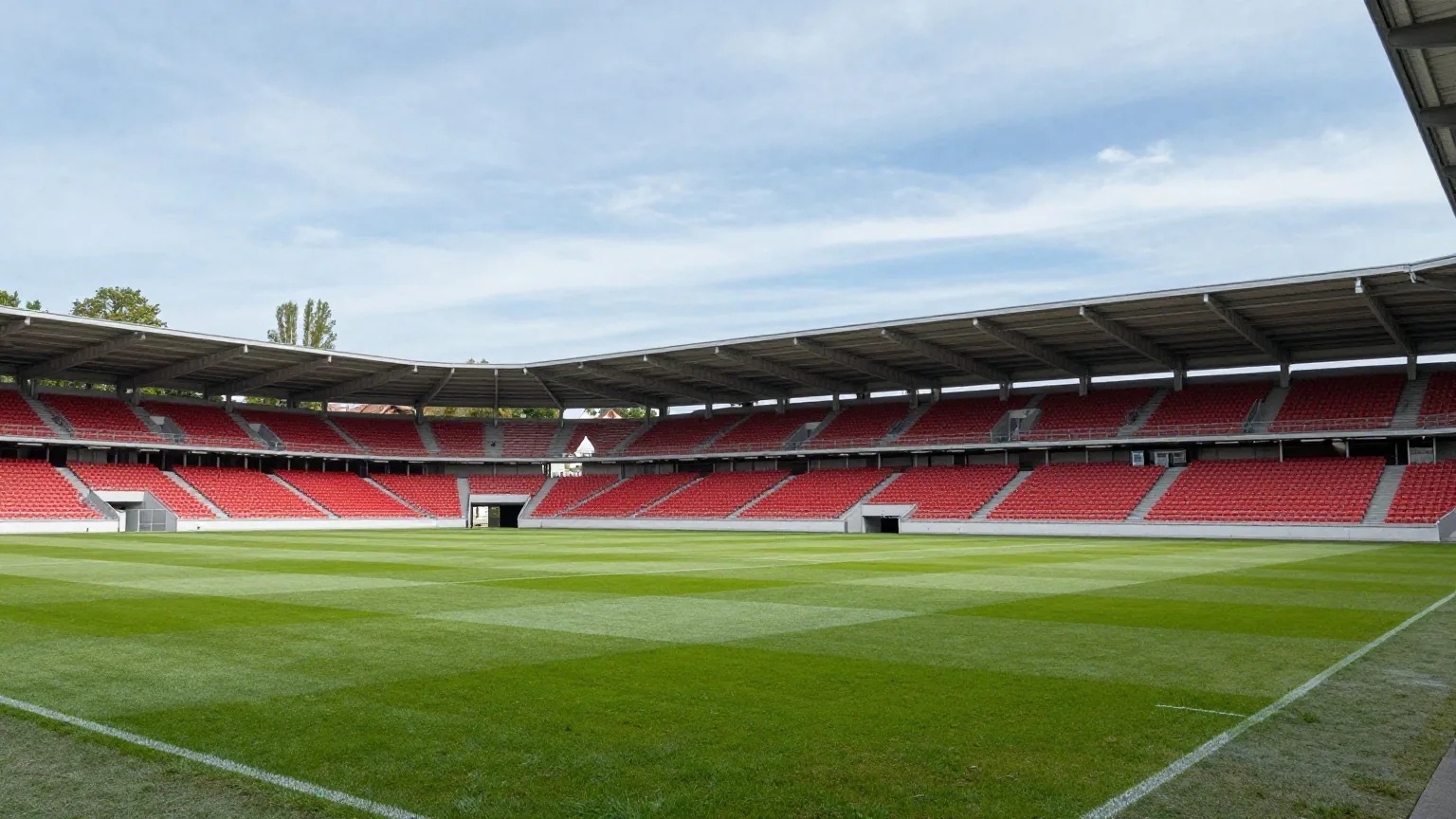 Kompaktes modernes Fußballstadion mit rotem Gestühl und durchgehendem Dach am Stadtrand unter blauem Himmel