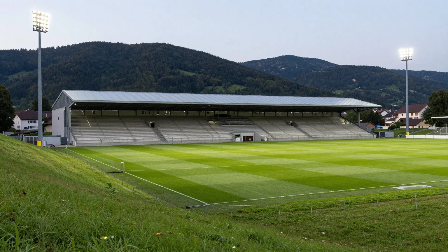 Modernes Fußballstadion mit hellgrauem Dach in der Abenddämmerung, dahinter bewaldete Hügel und leuchtendes Flutlicht