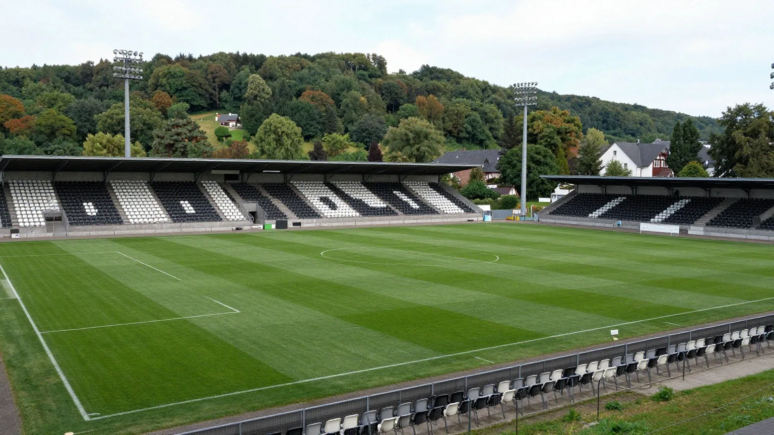 Kleines Fußballstadion am Waldrand mit schwarz-weißen Sitzen und vier Flutlichtmasten im Abendlicht