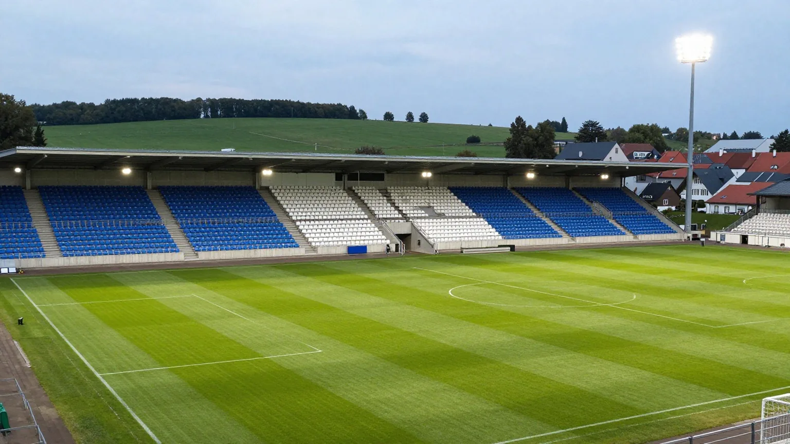 Kompaktes Fußballstadion in einer norddeutschen Kleinstadt in der Abenddämmerung mit blau-weißen Sitzen und eingeschaltetem Flutlicht