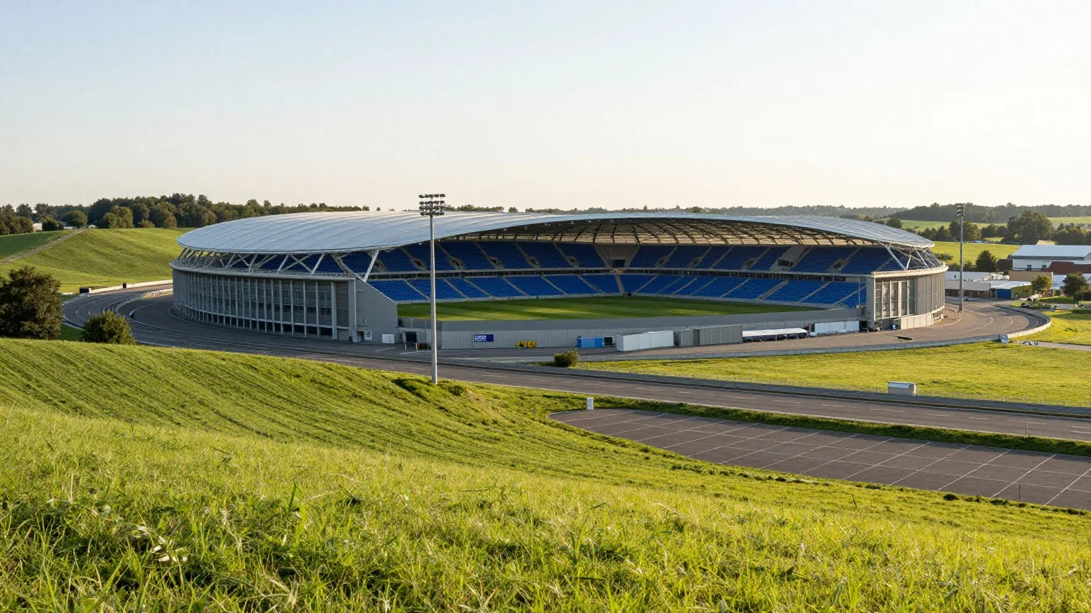 Großes modernes Stadion mit blauen Sitzen neben einer Autobahn in hügeliger grüner Landschaft im Abendlicht