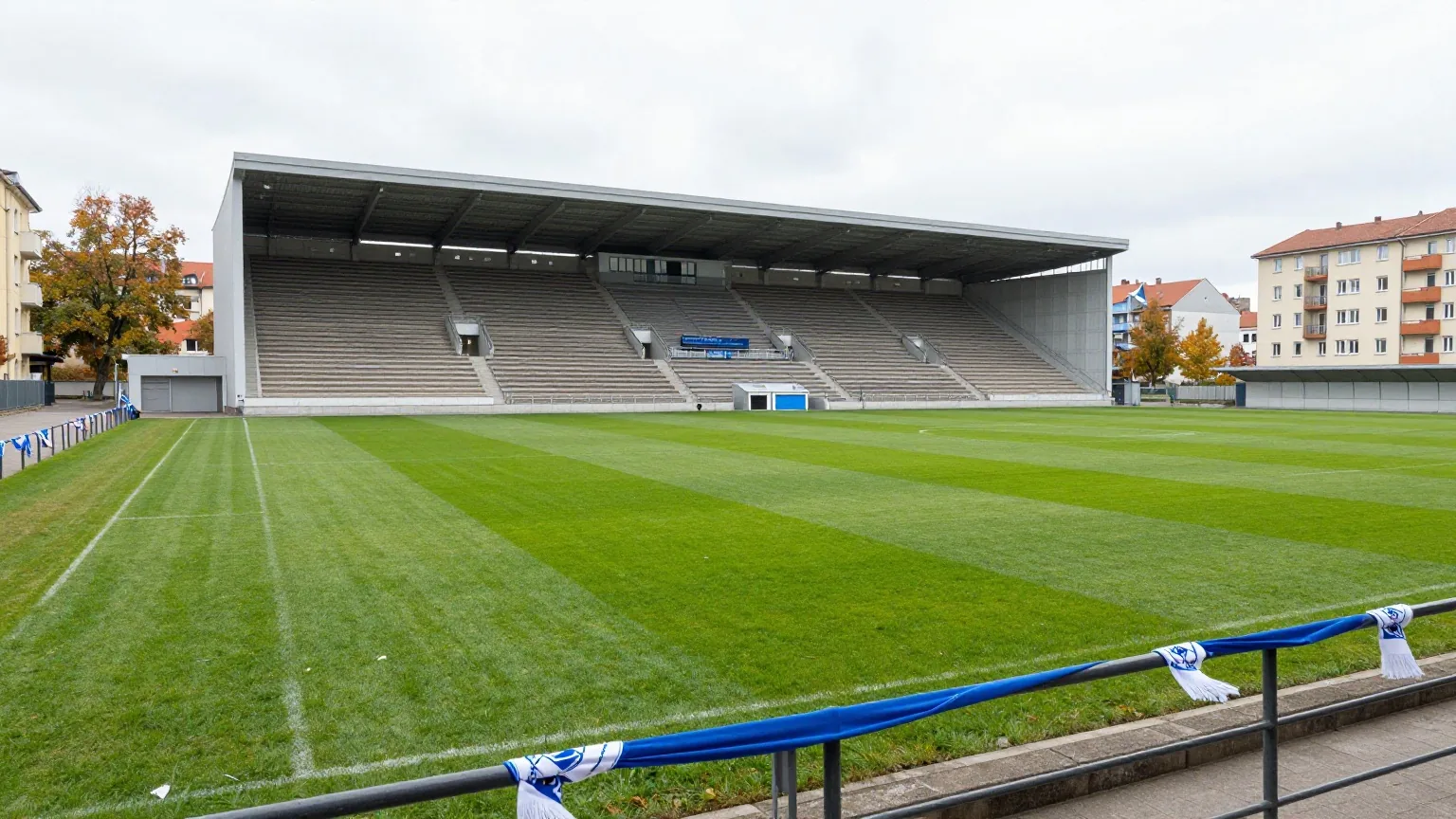 Altes Stadion mitten in der Stadt mit steilen Rängen, Wohnhäusern ringsum und hellblau-weißen Schals am Geländer