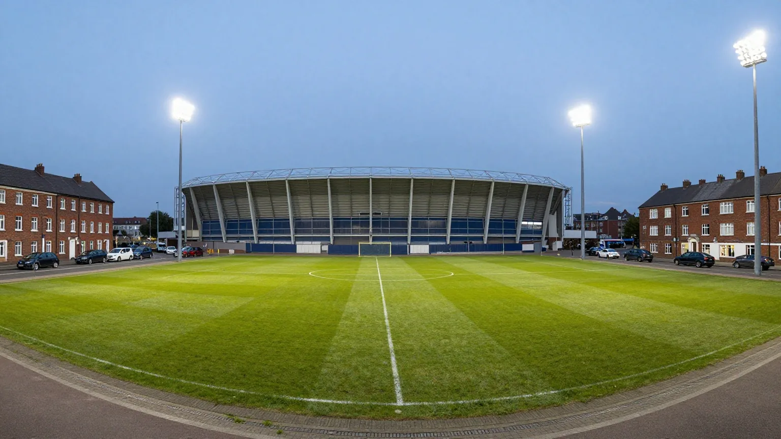 Kompaktes Fußballstadion zwischen Backsteinhäusern in einer Industriestadt am blauen Abendhimmel mit leuchtenden Flutlichtmasten